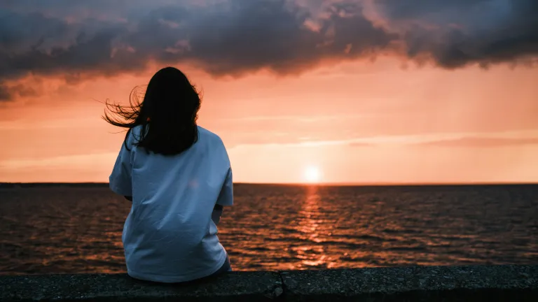 A woman sitting by a body of water watching the sunset.